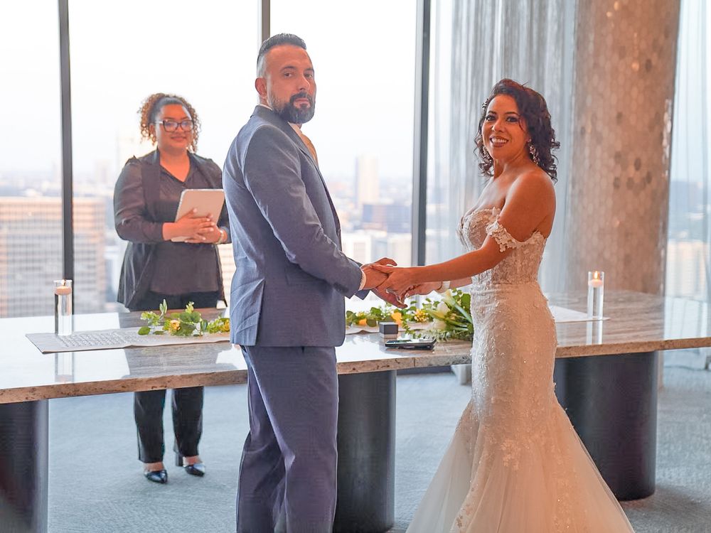 Bride and groom hold hands during indoor wedding ceremony with officiant and city skyline views.