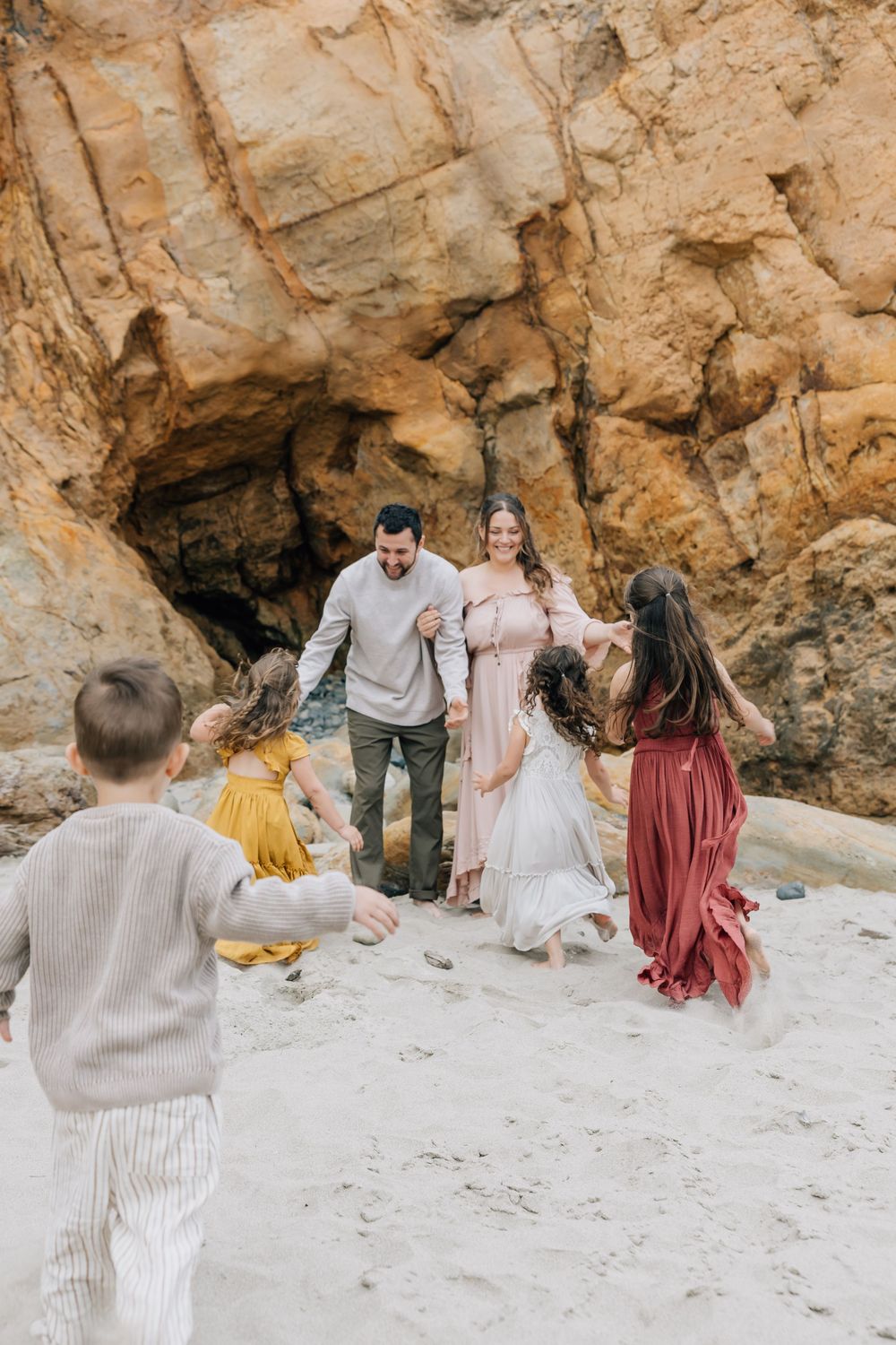 Family of five playing on sandy beach with rocky cliffs, parents smiling as children run toward them.