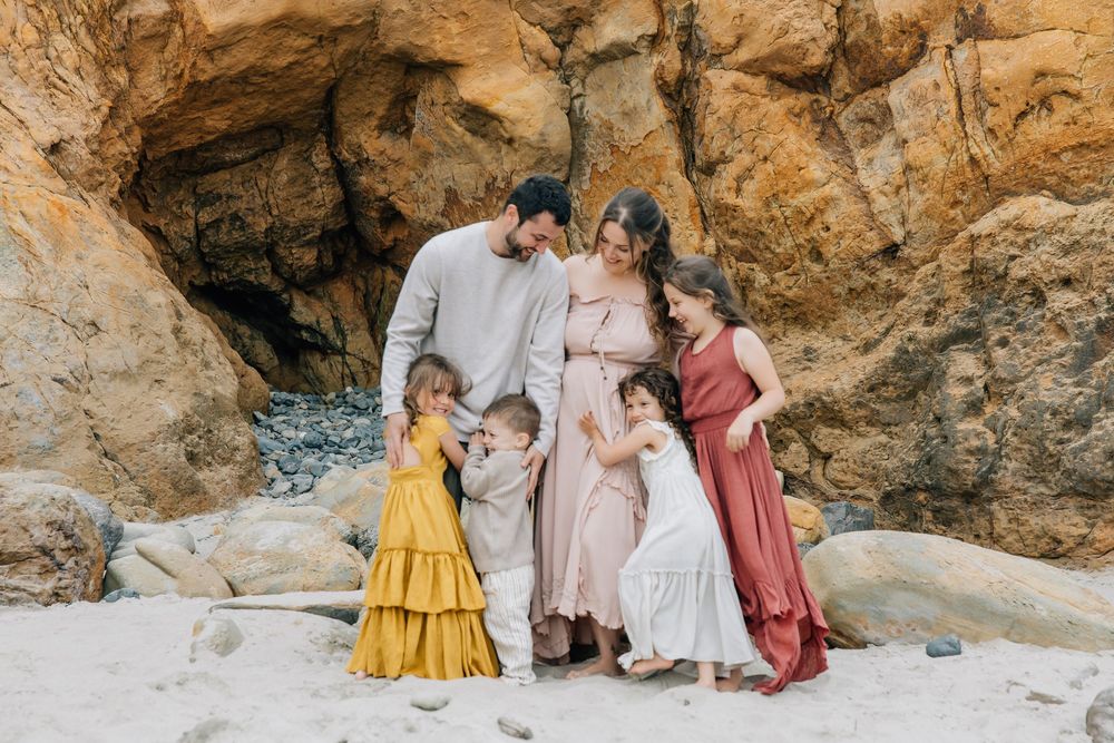A happy family of five shares a warm embrace near a rocky cave on a sandy beach.
