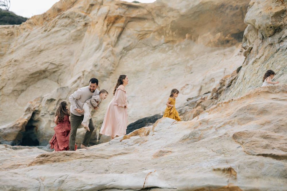 Family exploring rocky coastal cliffs, with a pregnant woman in a pink dress and children climbing the sandstone rocks.
