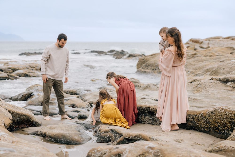 Family with young children exploring tide pools on a rocky beach, mother in pink dress holding a baby near the ocean.