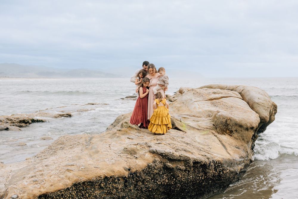 Family in colorful dresses standing on coastal rocks with ocean waves in the background on an overcast day.