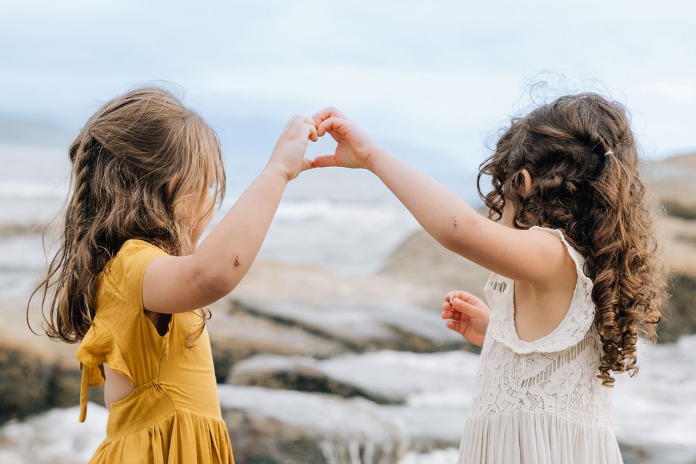 Two young girls making a heart shape with their hands at a rocky beach, wearing yellow and white dresses.