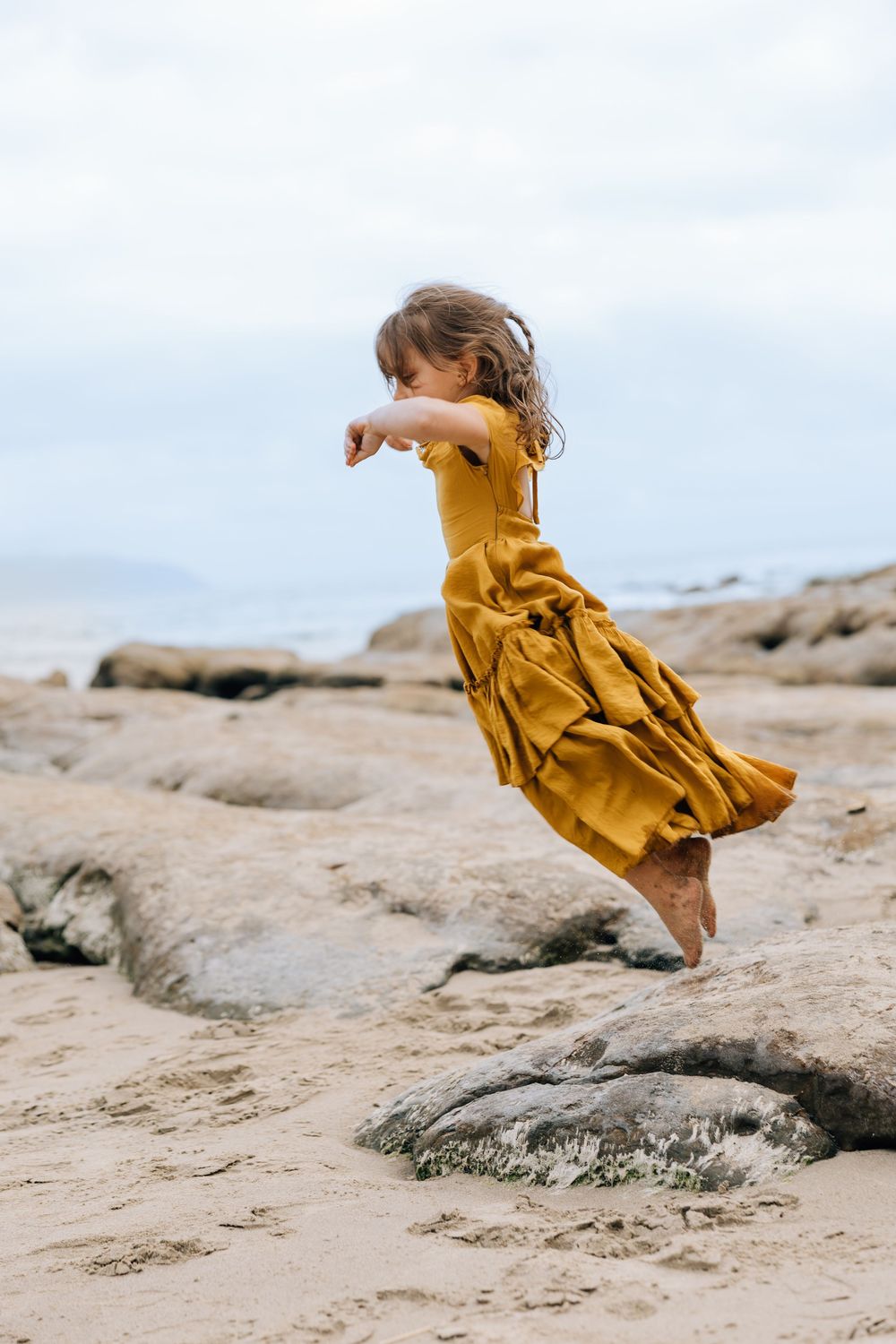 A young girl in a golden yellow dress leaps joyfully on rocky beach sand with ocean waves in the background.