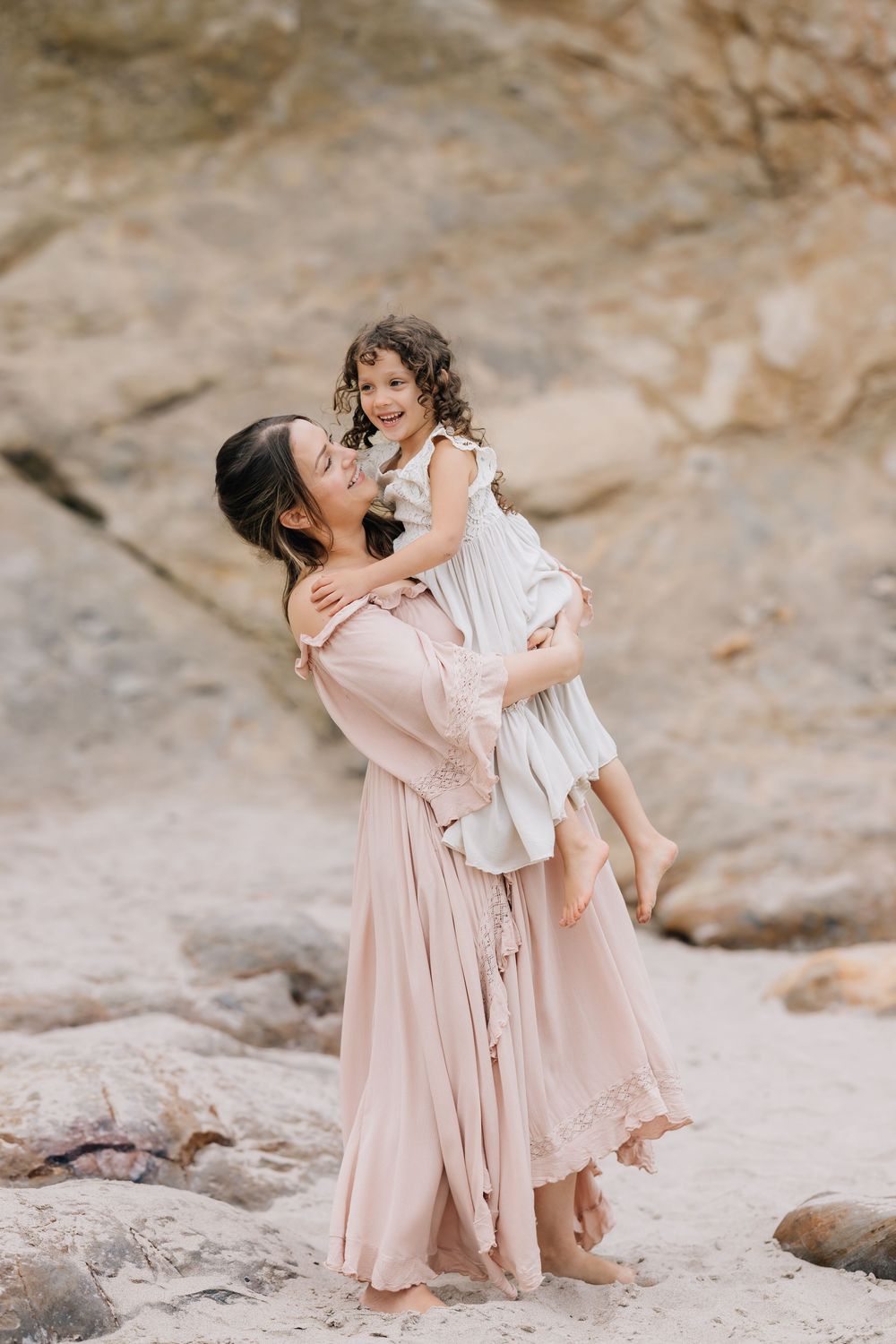 A joyful mother in a pink dress lifts her laughing curly-haired daughter near rocky cliffs at the beach.