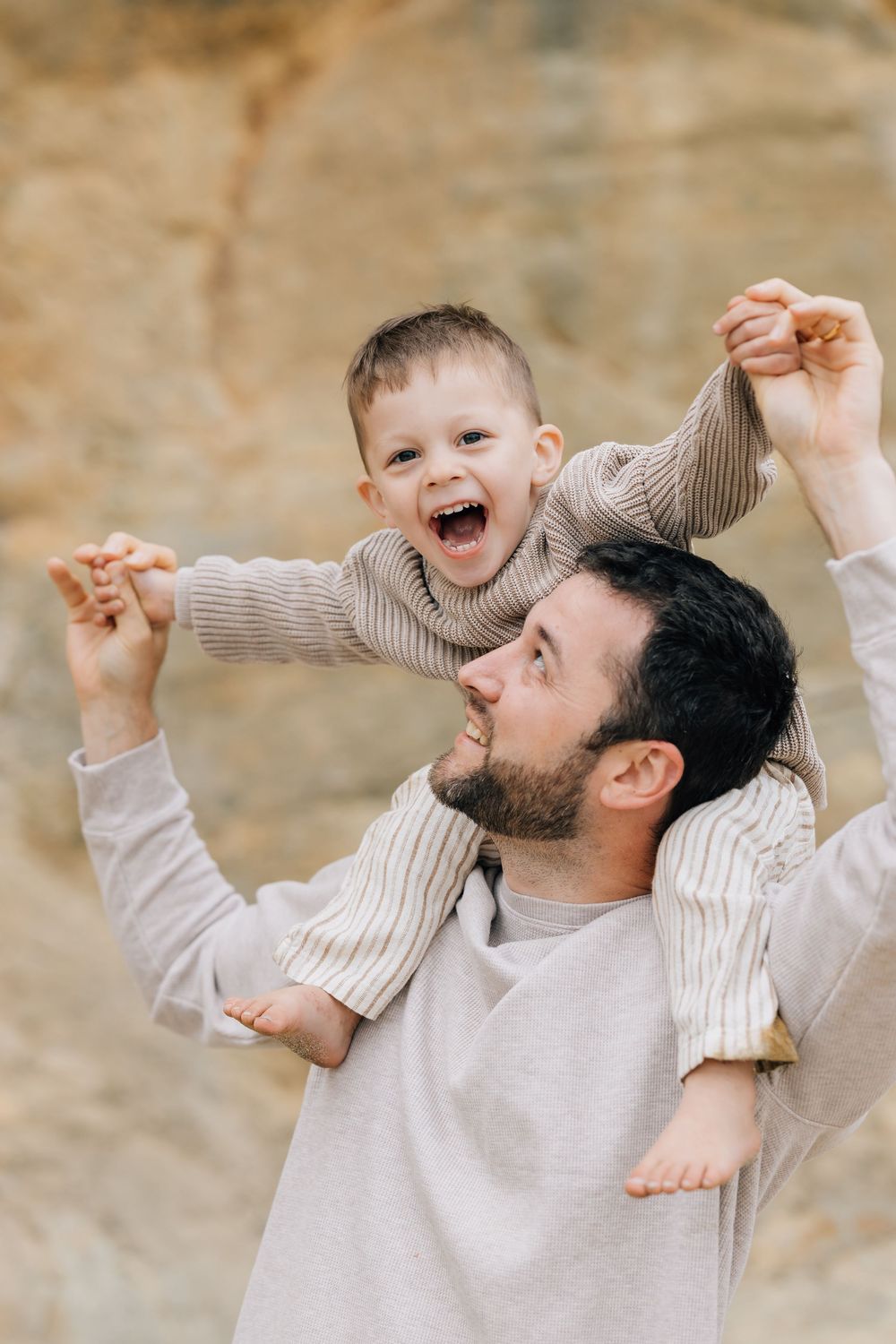 A bearded father lifts his laughing toddler son onto his shoulders outdoors near a rocky background.