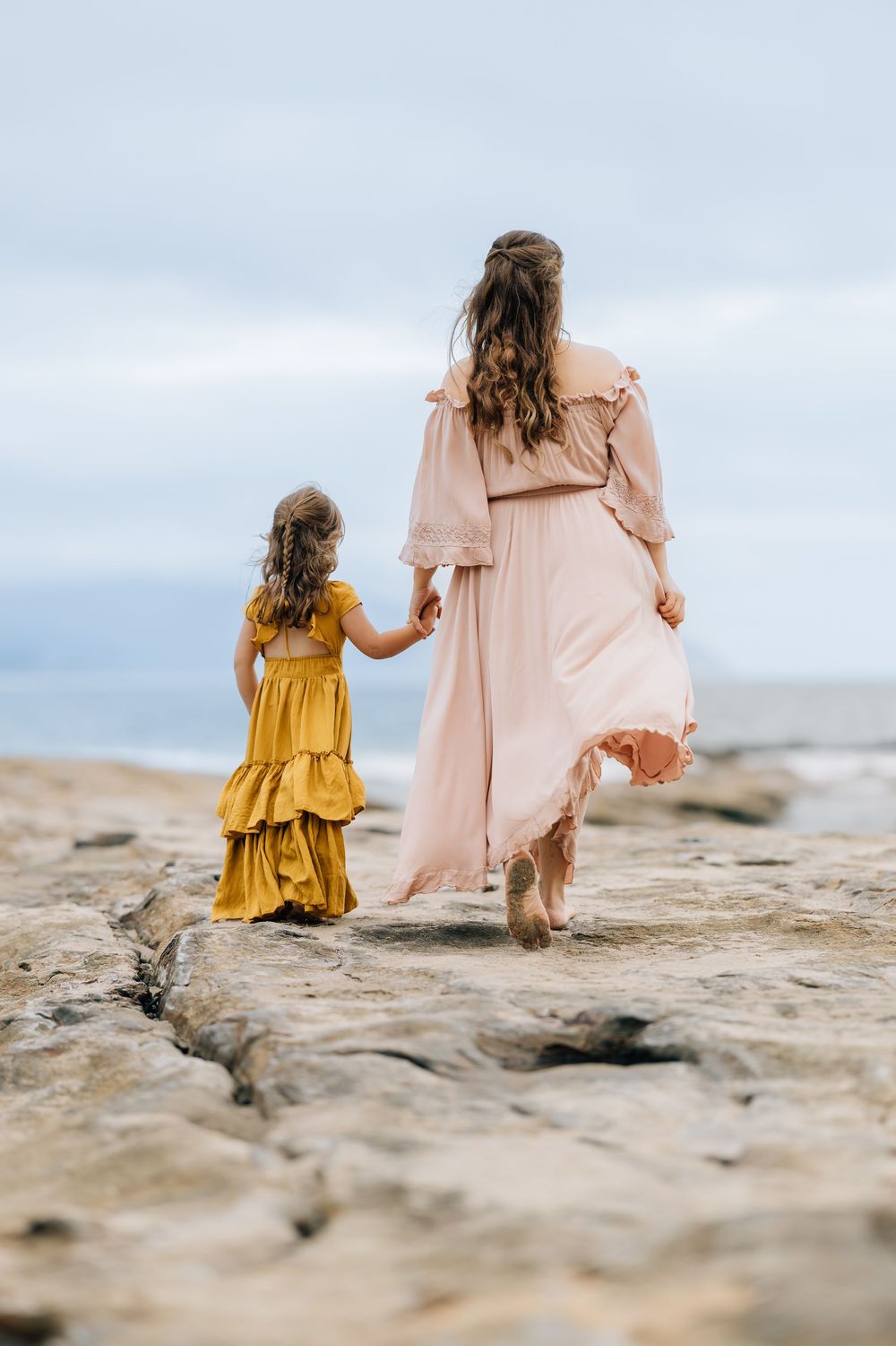 A mother and daughter in flowing dresses walk hand-in-hand on rocky coastal terrain toward the ocean.