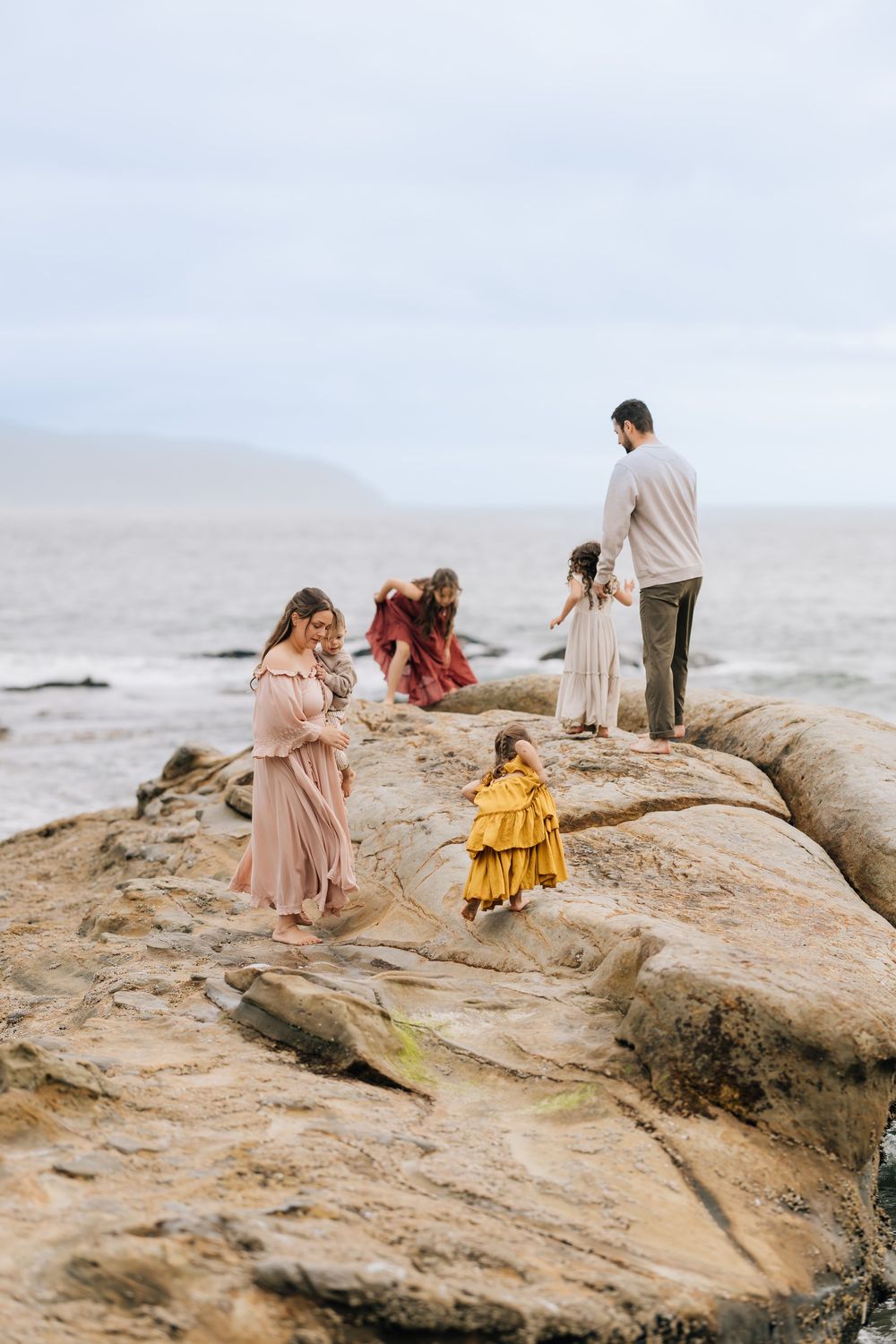 Family of six exploring rocky coastal shoreline, children in colorful dresses with parents overlooking the ocean.