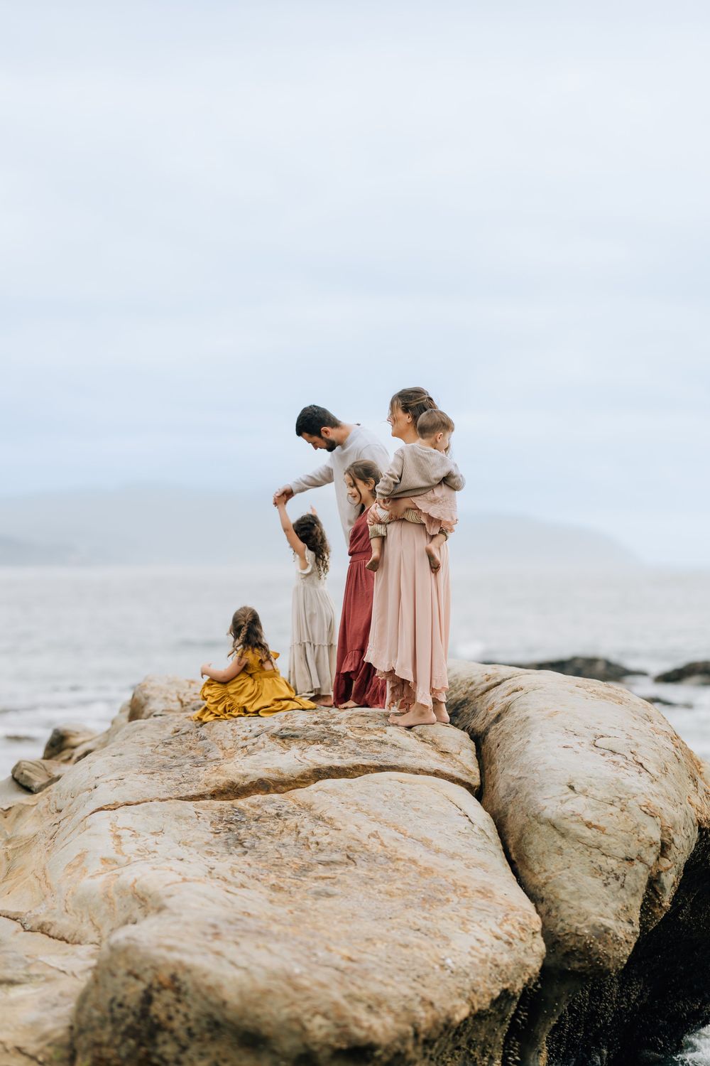 A family of five stands together on large coastal rocks overlooking the ocean on an overcast day.