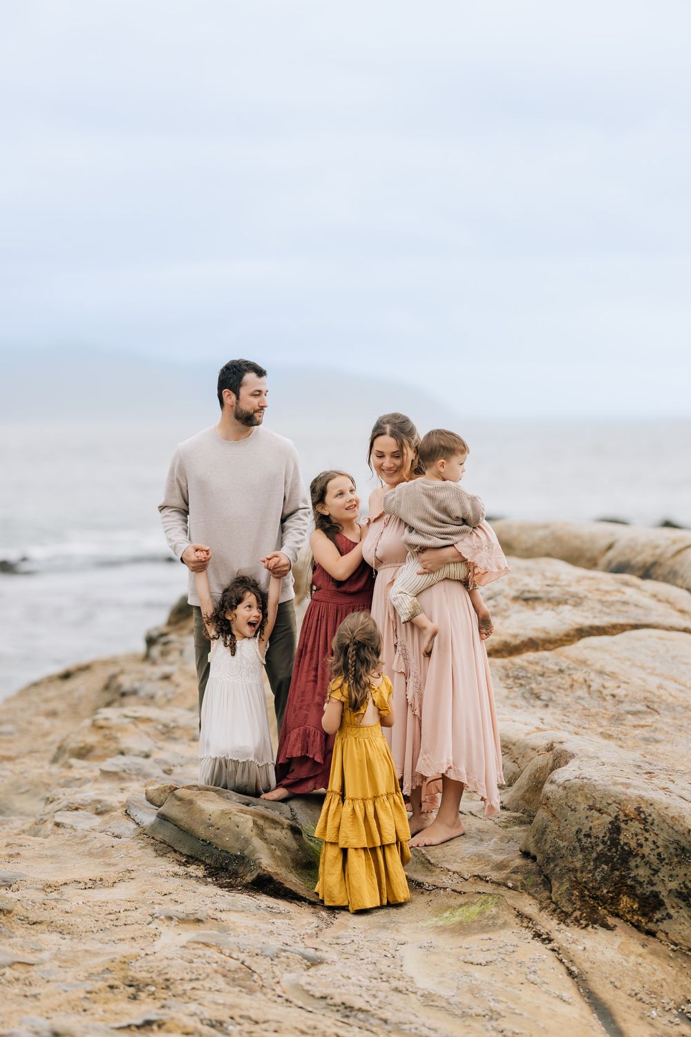Happy family of six posing on seaside rocks, mom in pink dress holding toddler with dad and three young children.