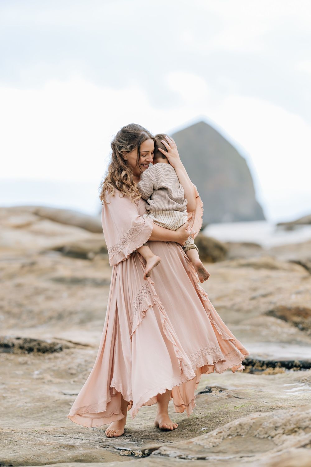A woman in a flowing pink boho dress holds a child on rocky beach with Haystack Rock in background.