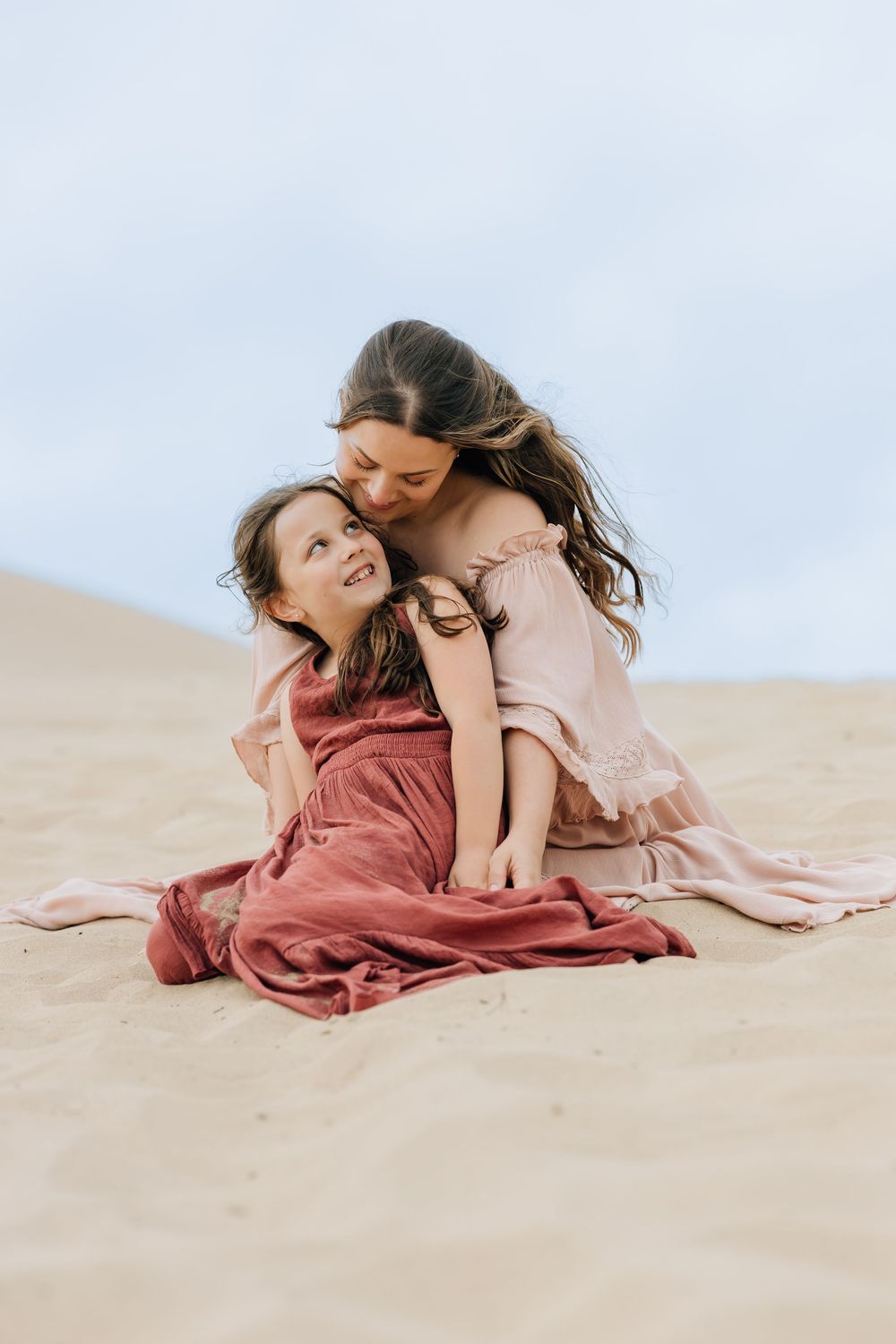 A mother in a pink dress embraces her daughter in a rose gown while sitting on sandy dunes.
