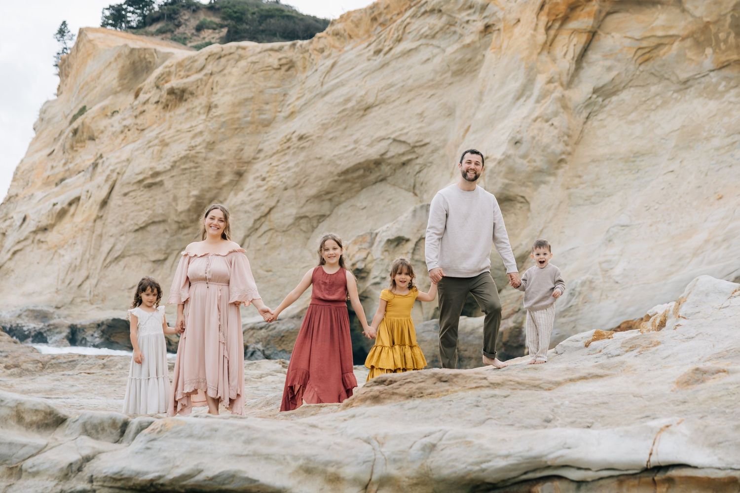 Family of six posing on rocky beach cliffs, wearing coordinated earth-tone outfits, holding hands outdoors.