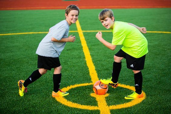 Two boys in soccer uniforms compete for a ball on a green turf field marked with yellow lines.