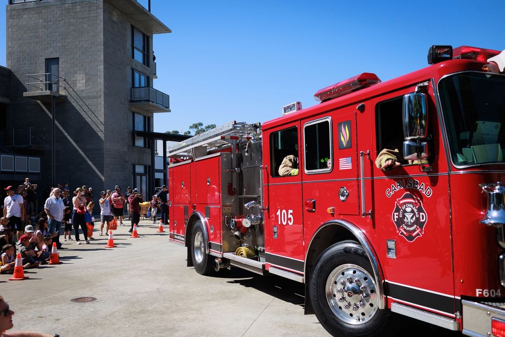 Red fire engine truck 105 parked at outdoor public event with crowd of spectators on sunny day.