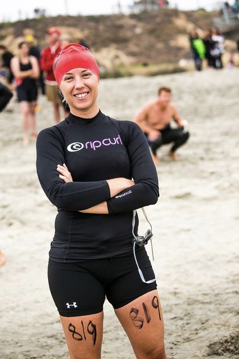 Woman in Rip Curl wetsuit and red swim cap smiling on beach with race number 819 written on her legs.