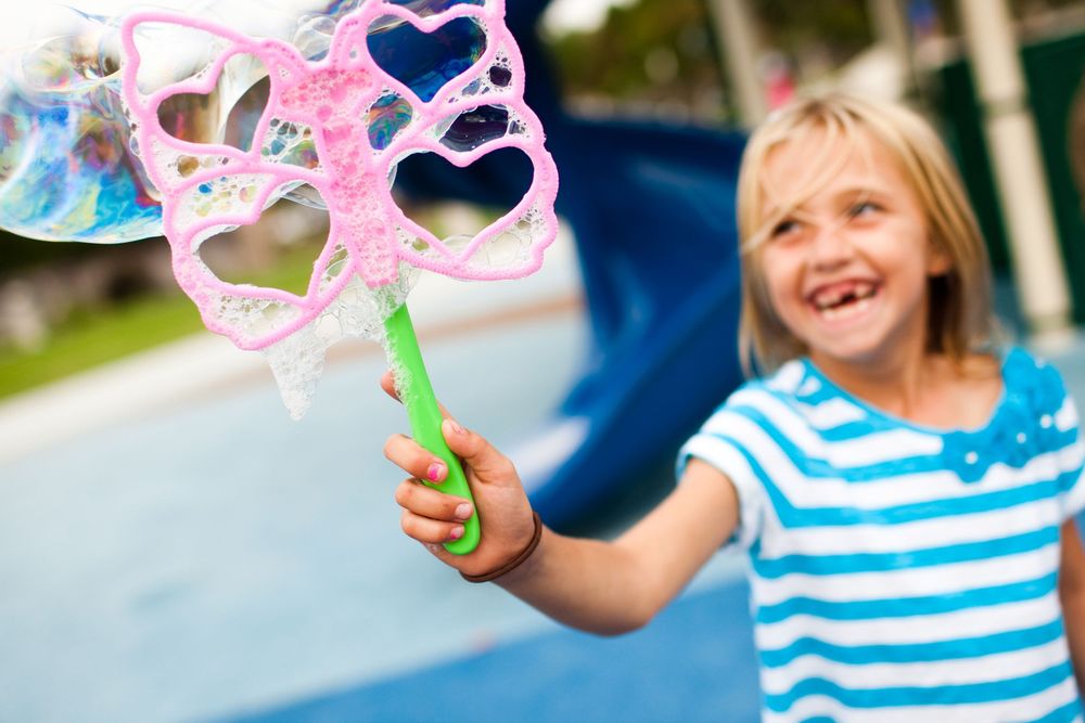 Young blonde girl smiling and holding a pink heart-shaped bubble wand at a playground.