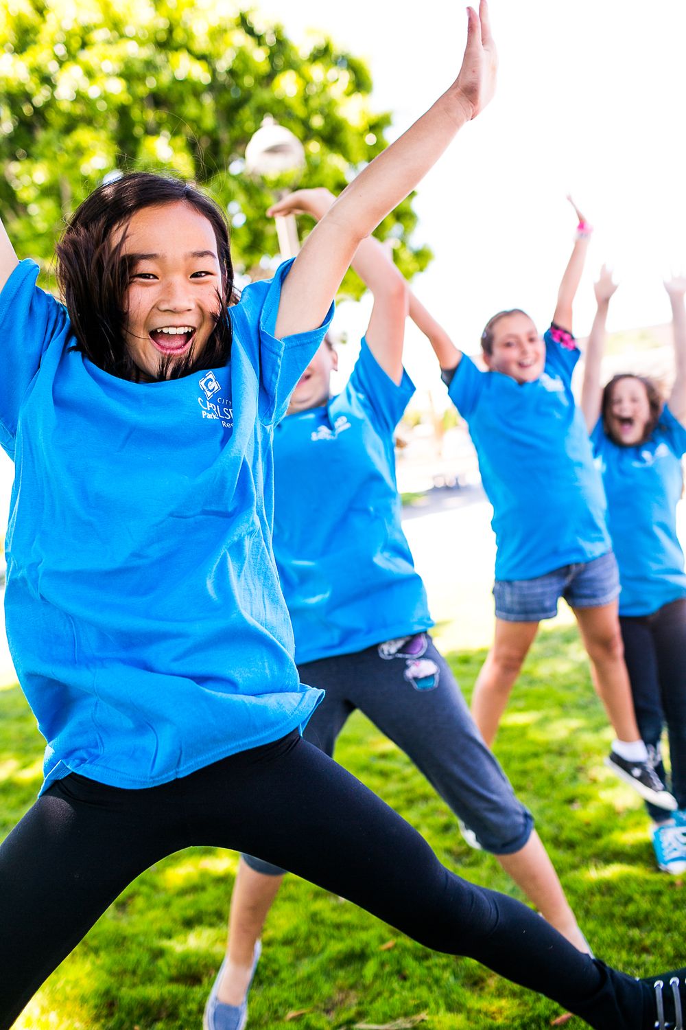 Girls in blue t-shirts joyfully dancing outdoors on green grass during a fun summer camp activity.