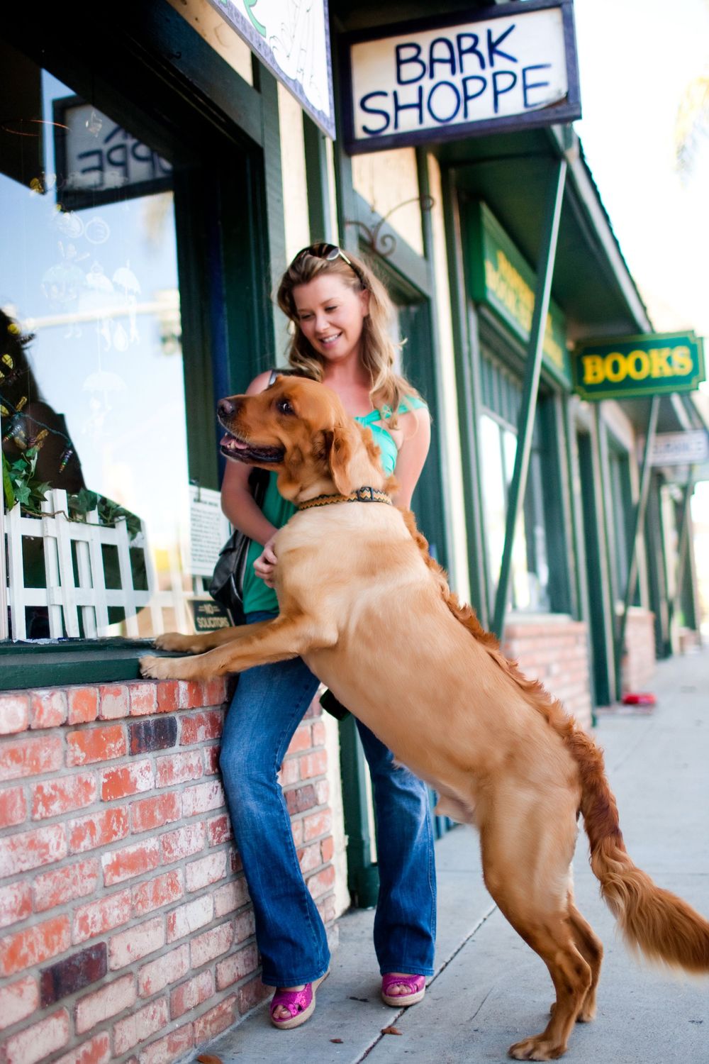 Woman hugging a golden retriever dog standing on hind legs outside Bark Shoppe pet store on a sunny day.