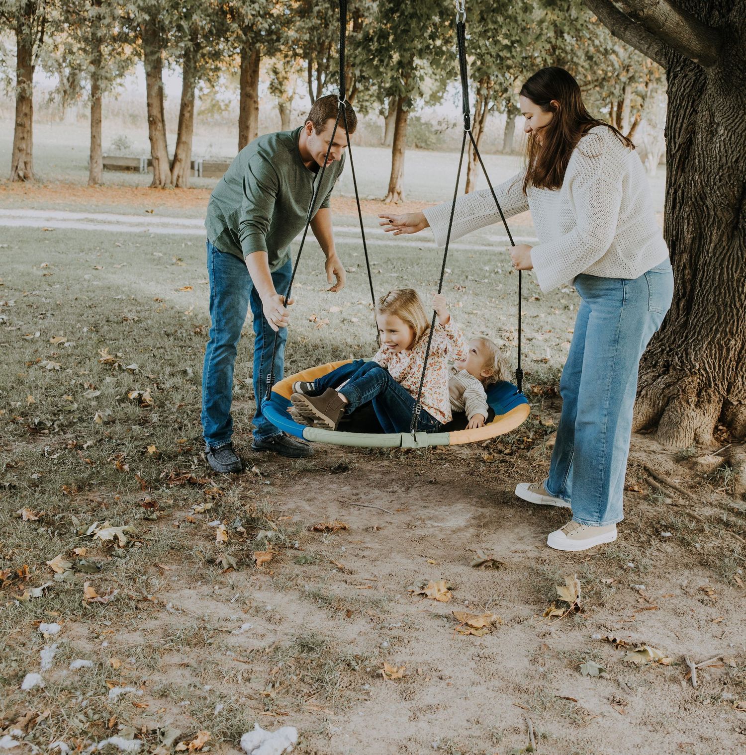 A family of four enjoys a sunny park day as two parents push their young children on a large round tree swing.
