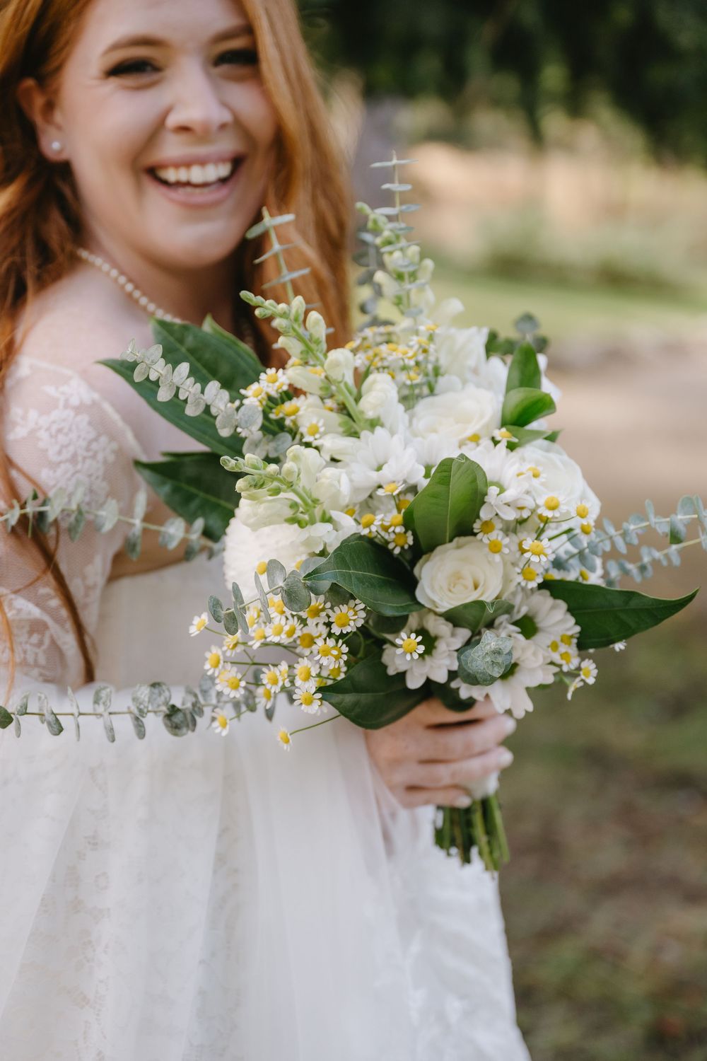 Smiling redhead bride in lace dress holds white bouquet with roses, daisies, and eucalyptus outdoors.