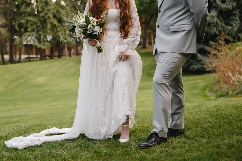 Bride in lace wedding gown holding white bouquet stands beside groom in gray suit on green lawn.