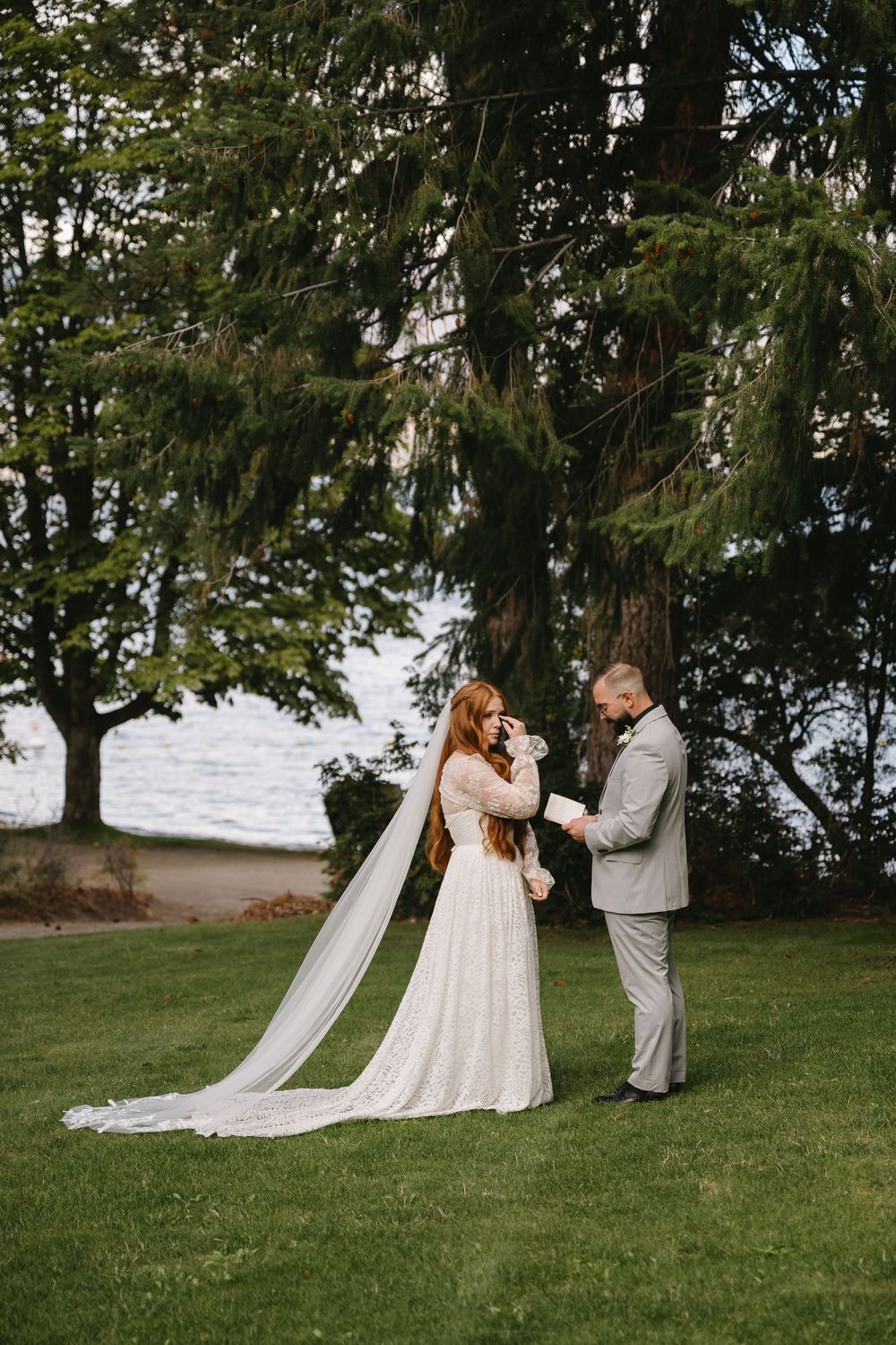 Bride and groom exchange vows outdoors on green lawn surrounded by tall evergreen trees near waterfront.