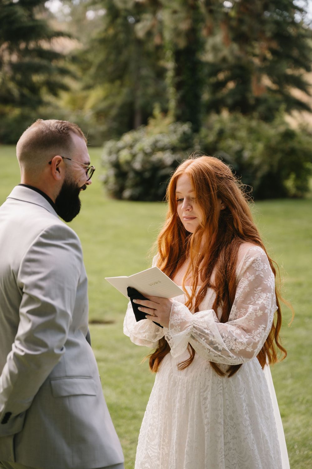 Red-haired bride in lace gown reads vows to bearded groom in gray suit during outdoor wedding ceremony.