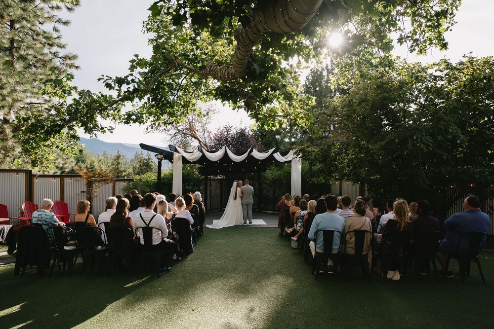 Outdoor wedding ceremony on green lawn with guests seated in rows facing couple at floral arch, surrounded by lush trees and mountains.