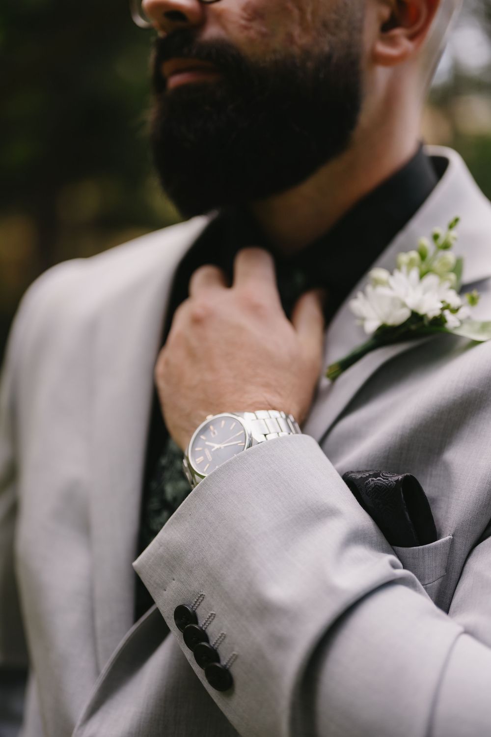 Bearded groom in gray suit adjusting black bow tie, wearing silver watch with white floral boutonniere.