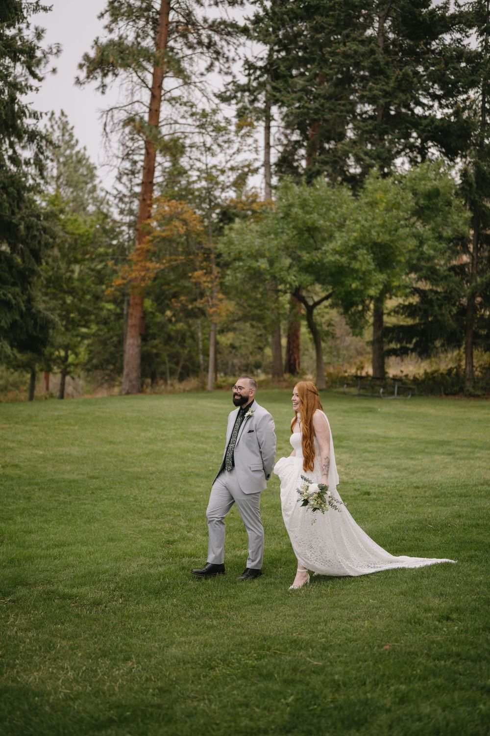 Bride in white lace gown and groom in gray suit walk hand in hand on green lawn surrounded by tall pine trees.