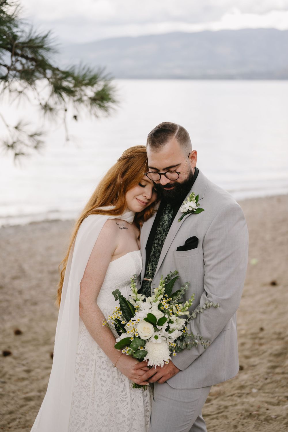 Bride and groom embrace on sandy beach, she holds white bouquet, he wears gray suit, mountains visible in background.