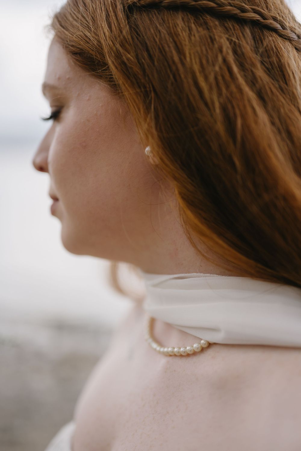 Close-up profile of a redhead bride wearing a pearl necklace and braided hair accessory with a white ribbon.