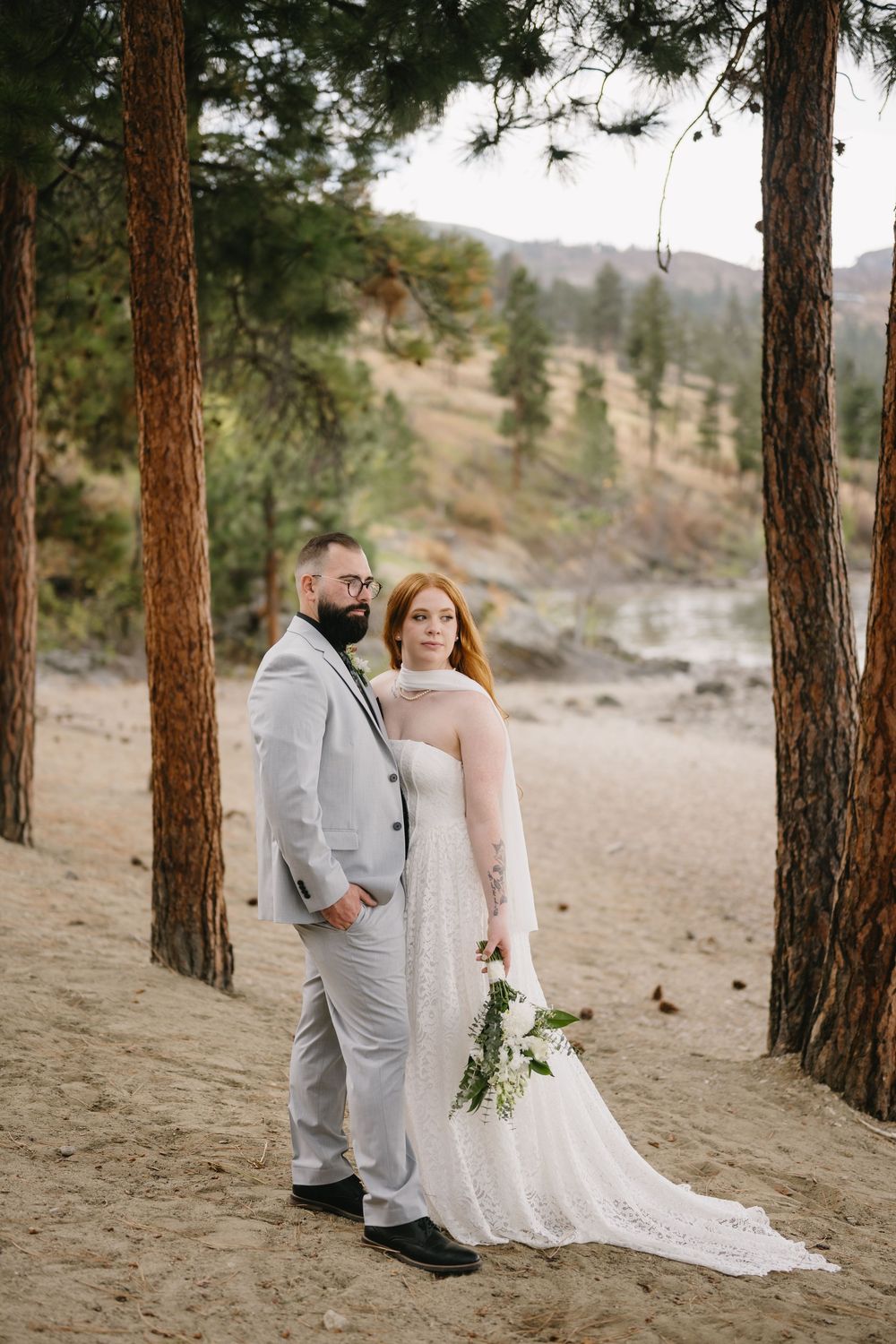 Bride and groom portrait in a pine forest, she in lace gown holding greenery bouquet, he in grey suit.