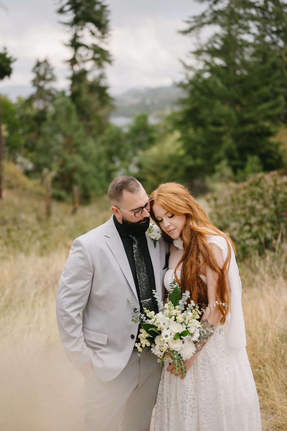 Bride and groom embrace in a forest meadow, she holds white bouquet in lace gown, he wears a gray suit.