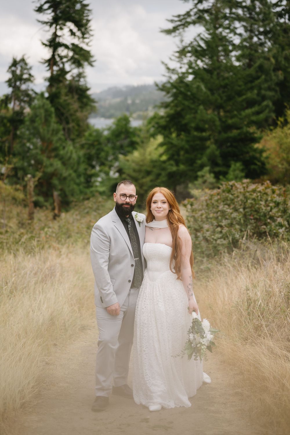 Bride and groom posing on a dirt path surrounded by tall trees and golden grass on their wedding day.