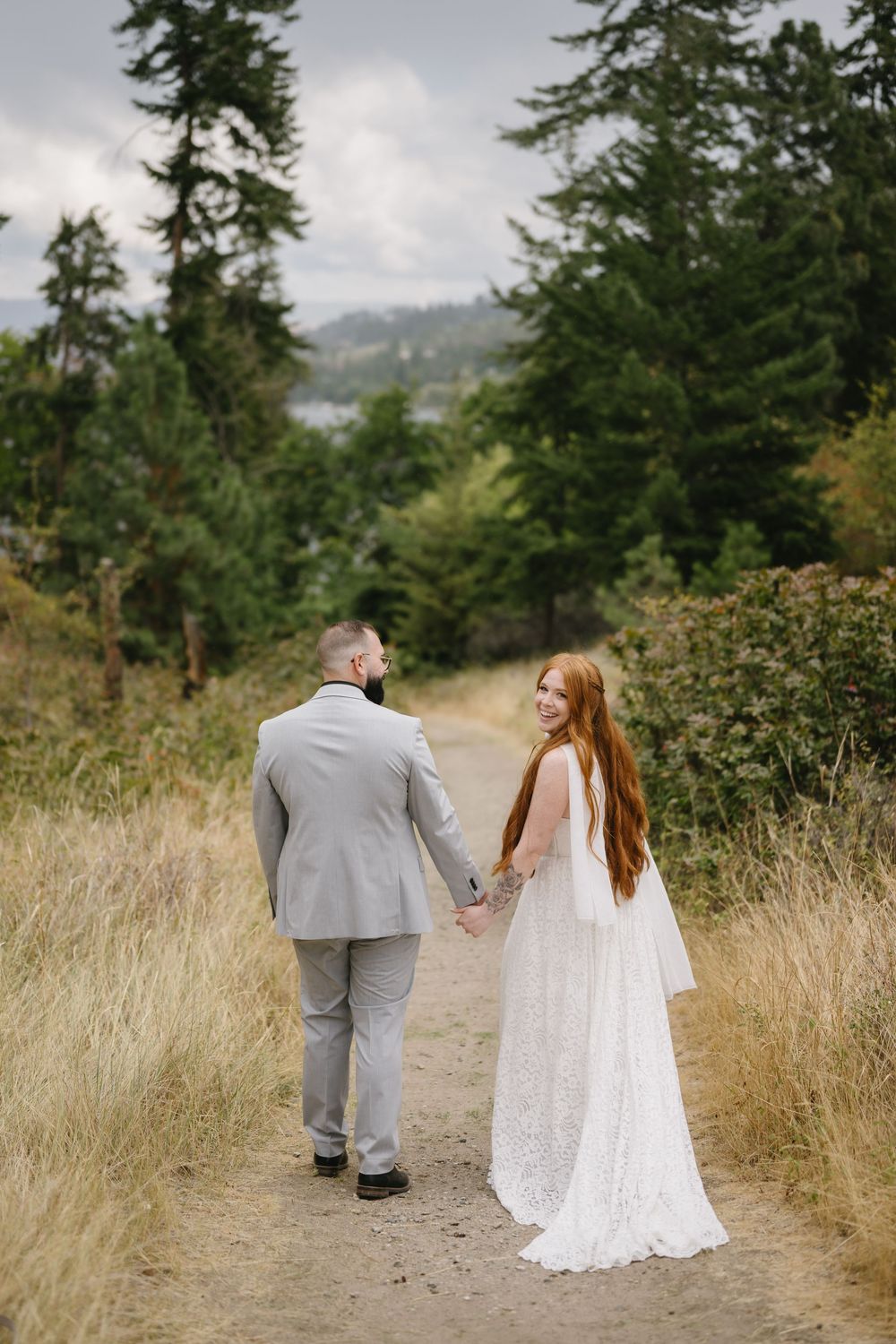 A bride and groom walk hand-in-hand down a forest path, she in a white lace gown, he in a gray suit.