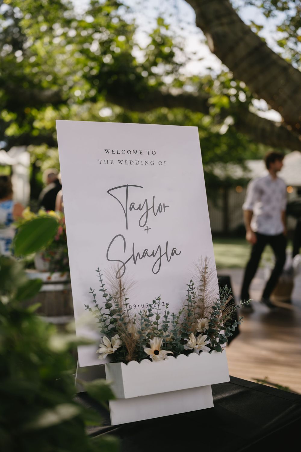 Elegant outdoor wedding welcome sign for Taylor and Shayla, adorned with white flowers and greenery.