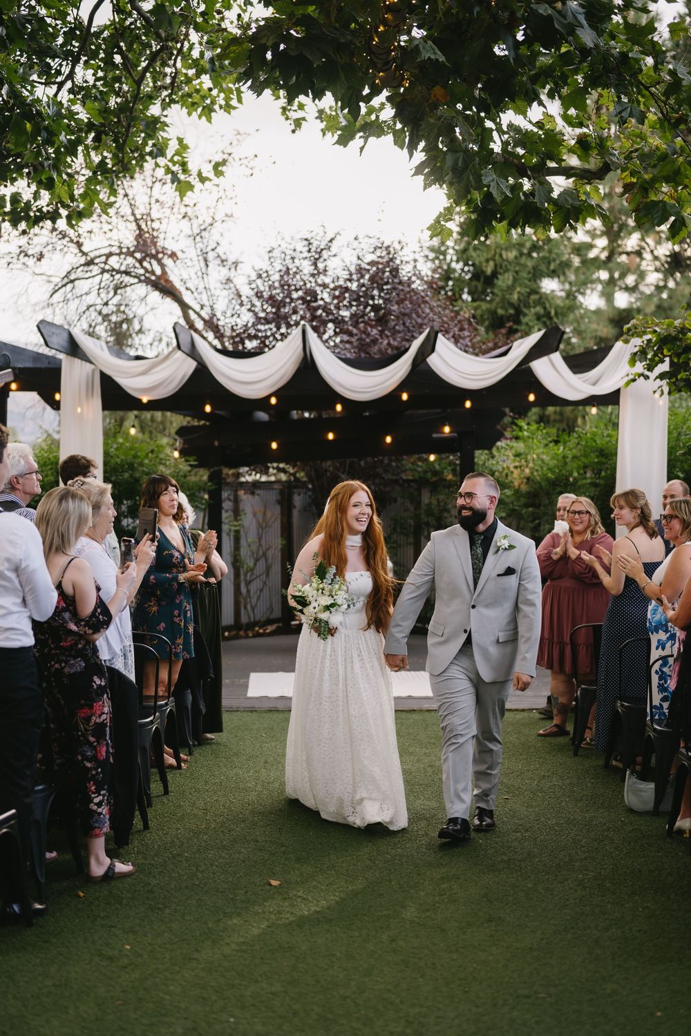 Newlyweds walk hand-in-hand down the aisle at an outdoor garden wedding ceremony with cheering guests.