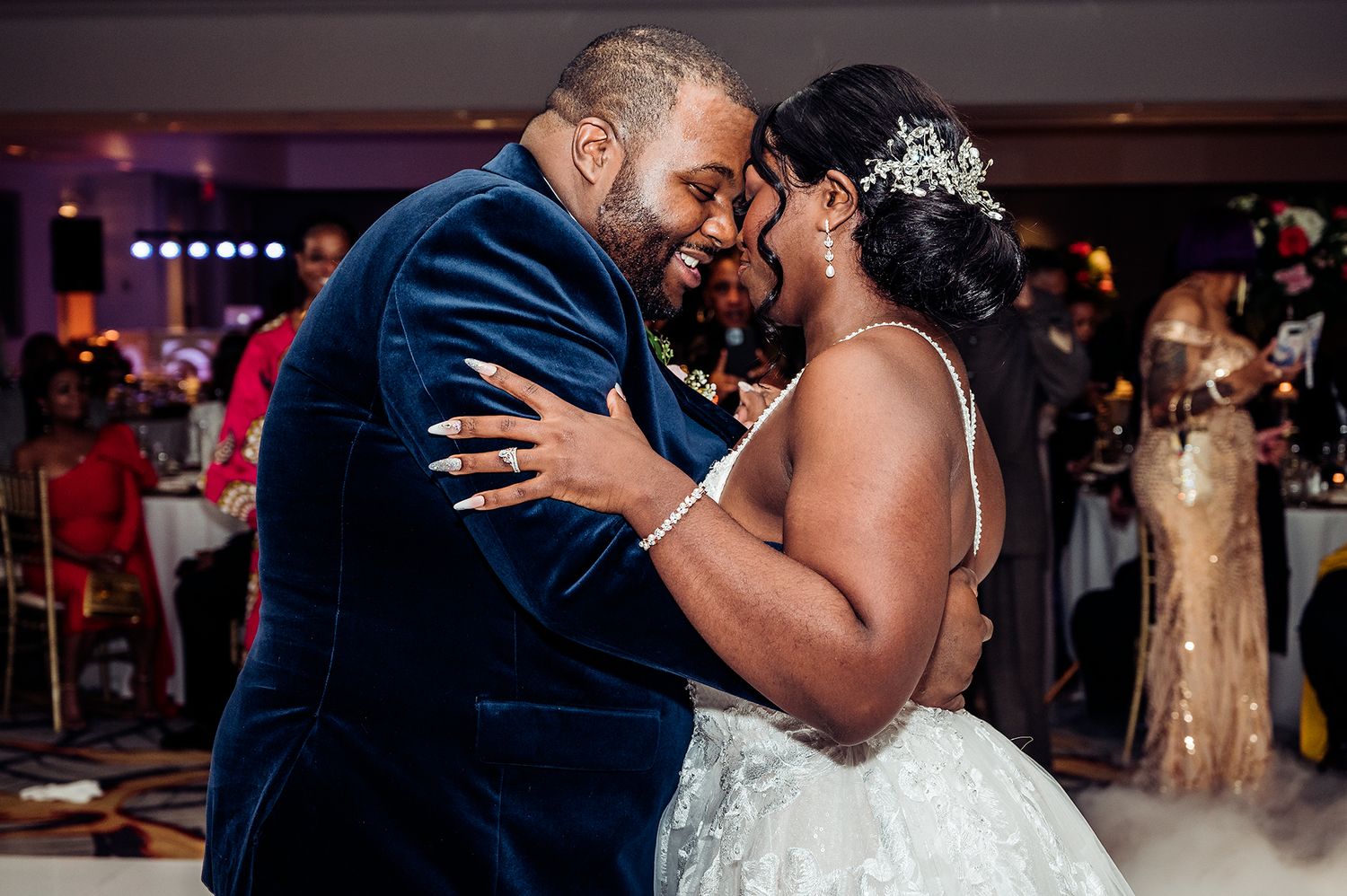 Bride and groom share romantic first dance at wedding reception, groom in blue velvet suit, bride in white lace gown.