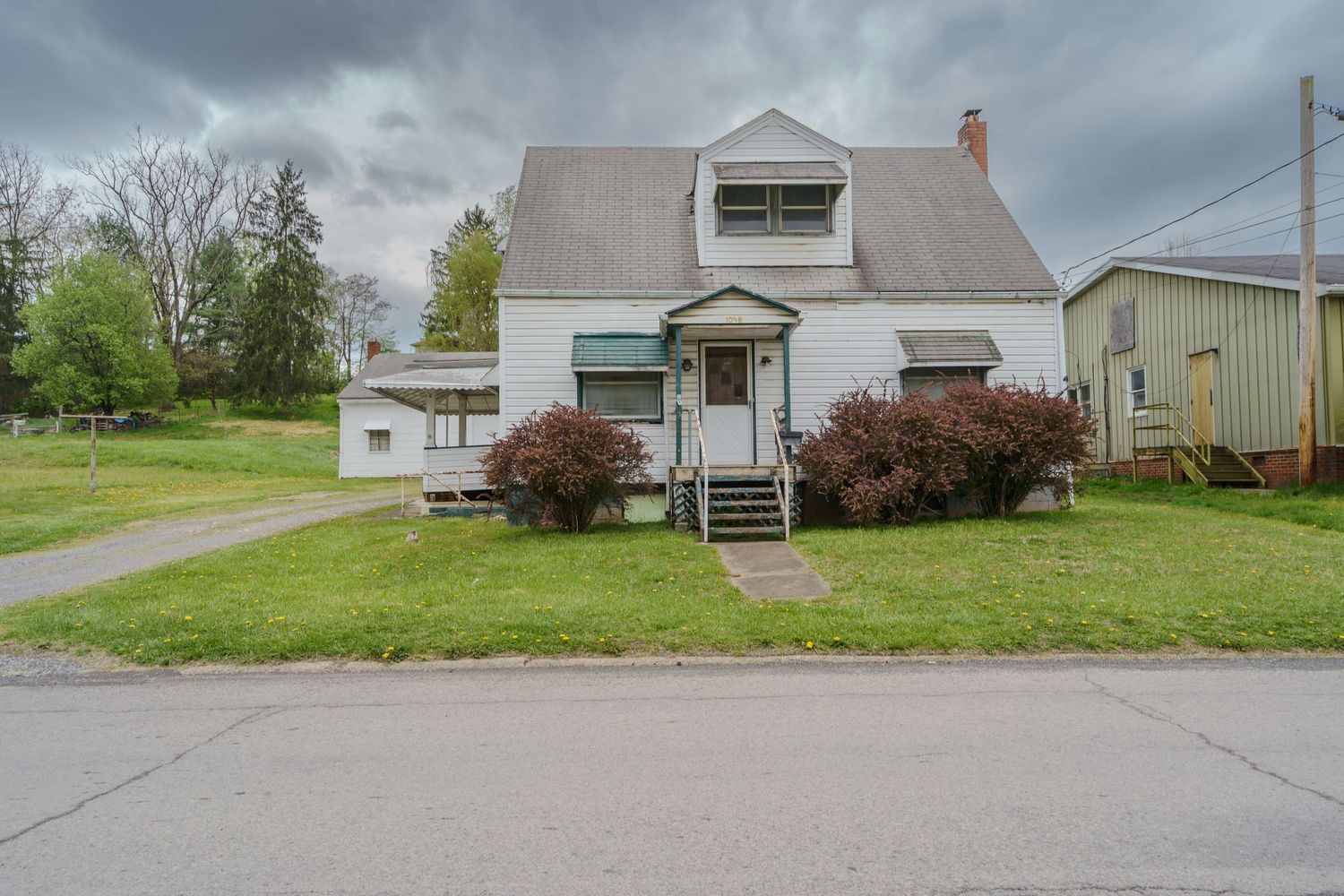 White cape cod home with dormers, red bushes, and green lawn on a quiet residential street under cloudy skies.