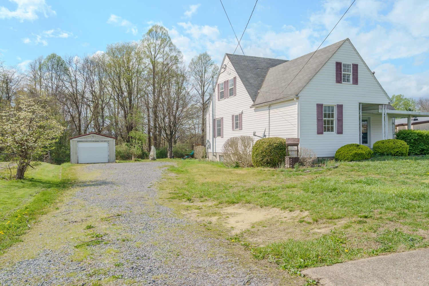 White colonial home with detached garage, gravel driveway, and lush green lawn surrounded by trees in spring.