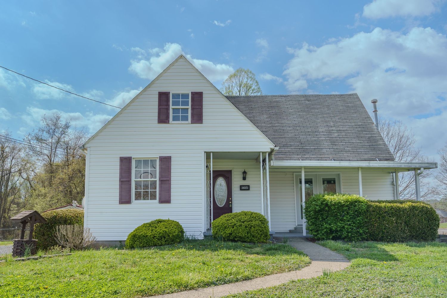 White Cape Cod home with dark shutters, covered porch, lush green lawn, and mature shrubs on a sunny day.