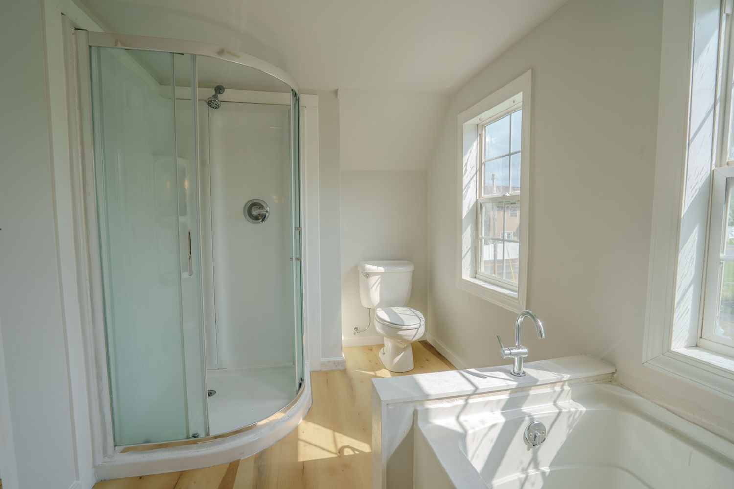 Bright bathroom featuring a glass corner shower, freestanding soaking tub, toilet, and hardwood floors with natural light.