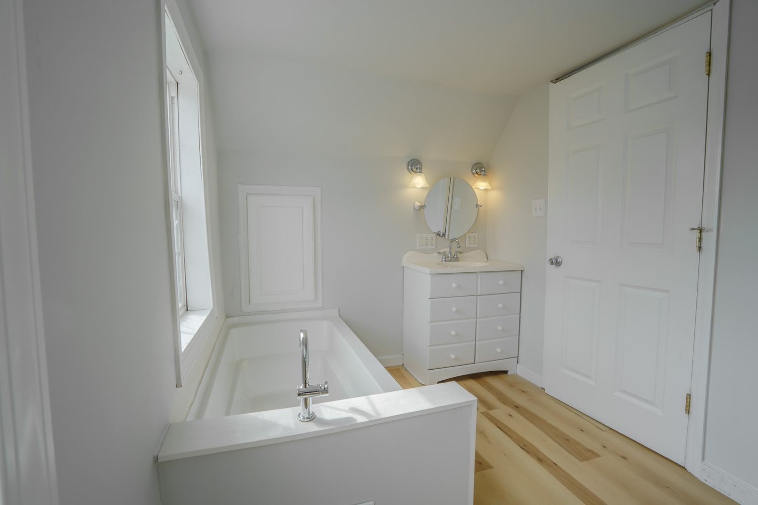 Modern white bathroom with freestanding soaking tub, wood floors, round mirror vanity, and bright natural light from window.