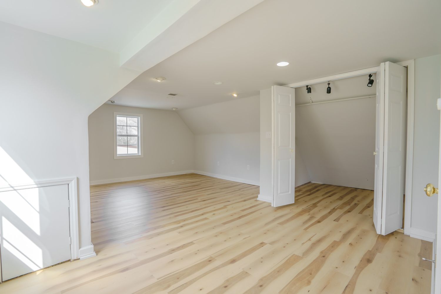 Bright open attic bonus room with light hardwood floors, white walls, columns, track lighting, and a bifold closet door.