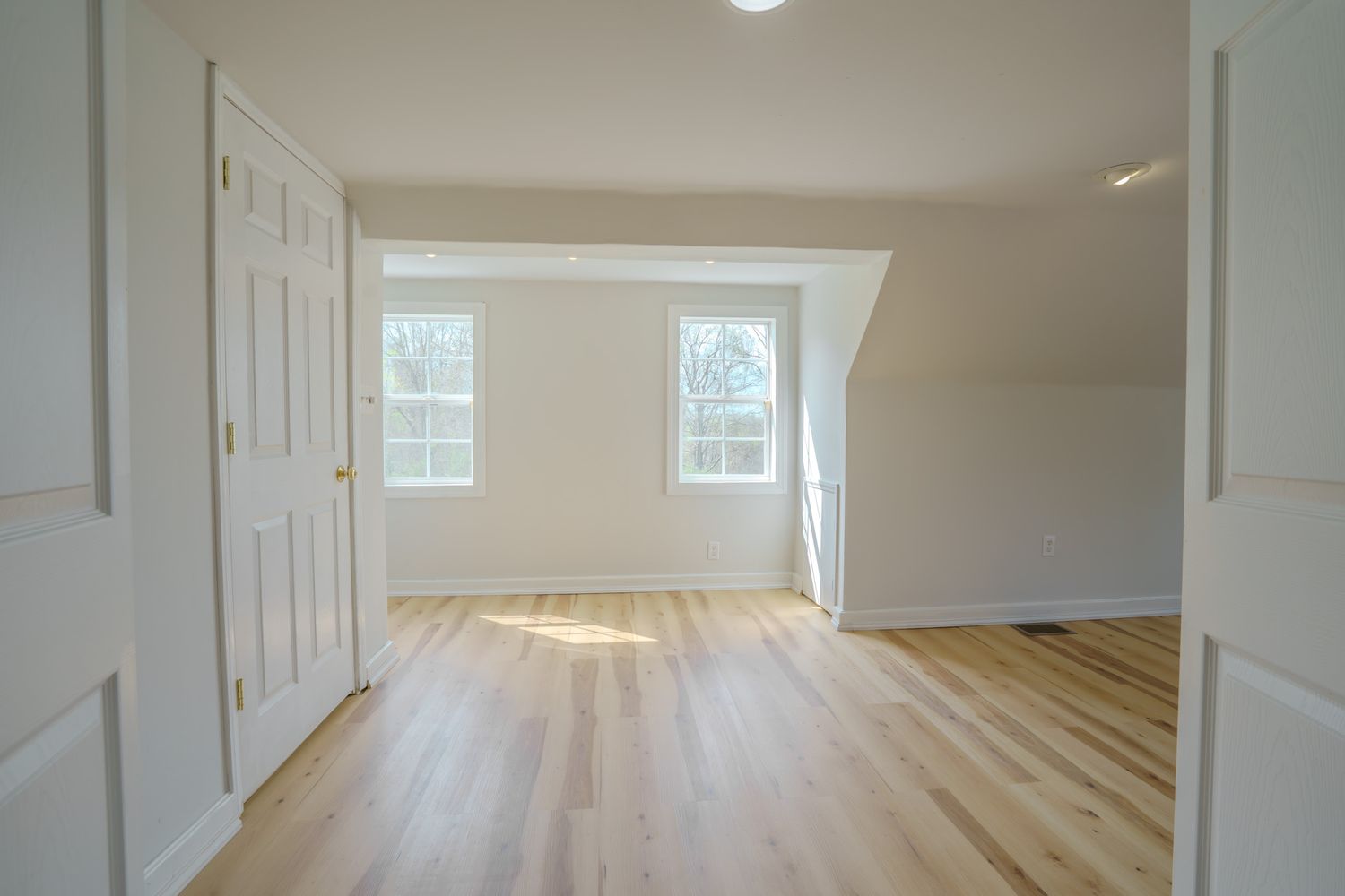 Empty room with light hardwood floors, white walls, sloped ceiling, and two windows letting in natural light.