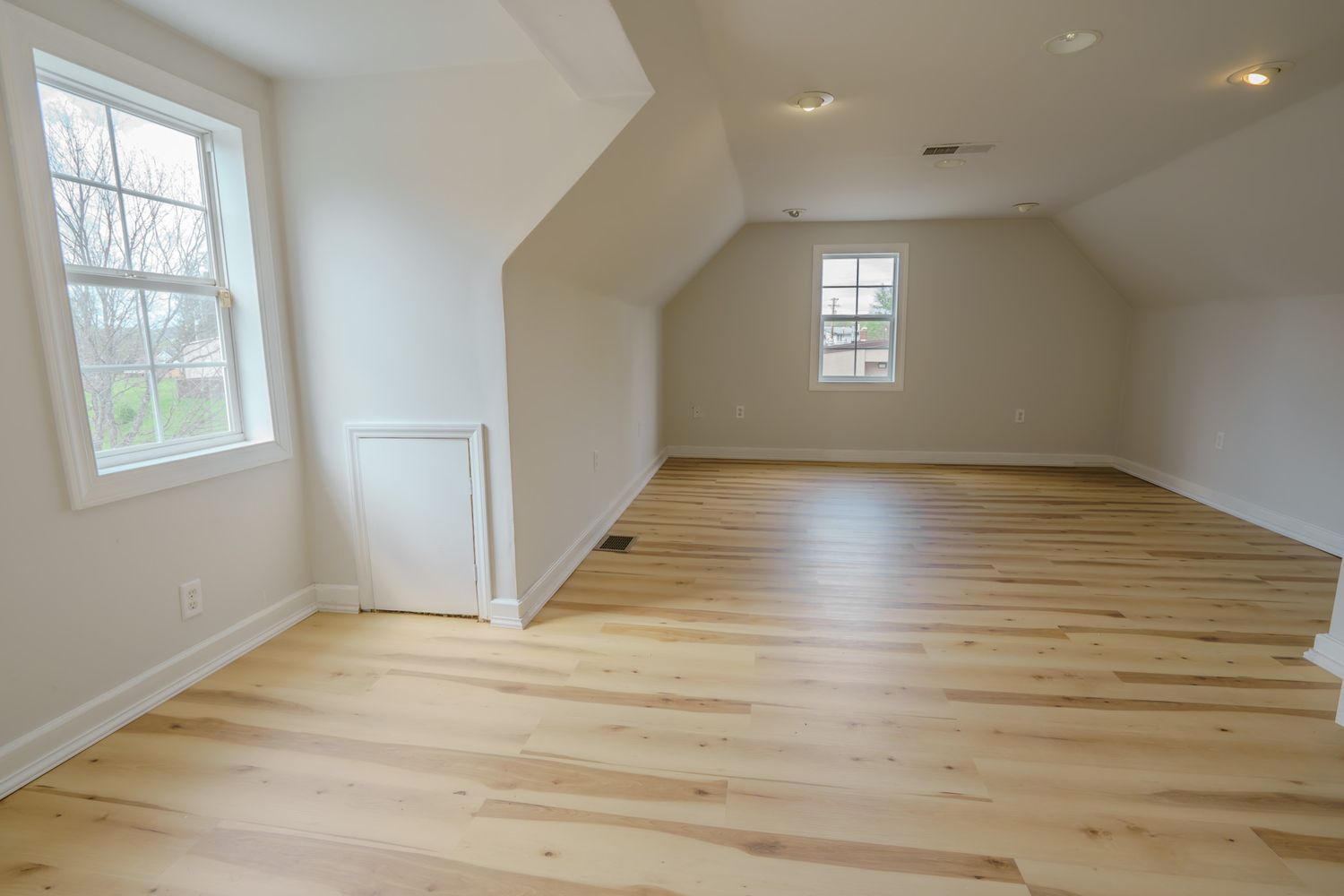 Bright attic room with vaulted ceiling, light hardwood floors, white walls, and two windows with natural light.