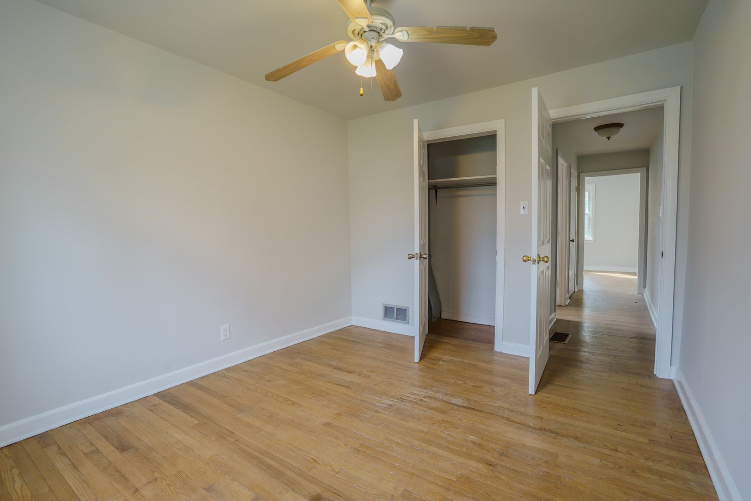 Bright bedroom with hardwood floors, ceiling fan, open closet, and white trim leading to a hallway.