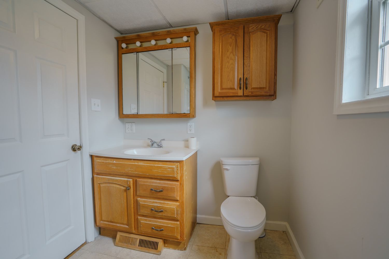 Clean bathroom with oak vanity, medicine cabinet, white toilet, and natural light from window on tile floor.